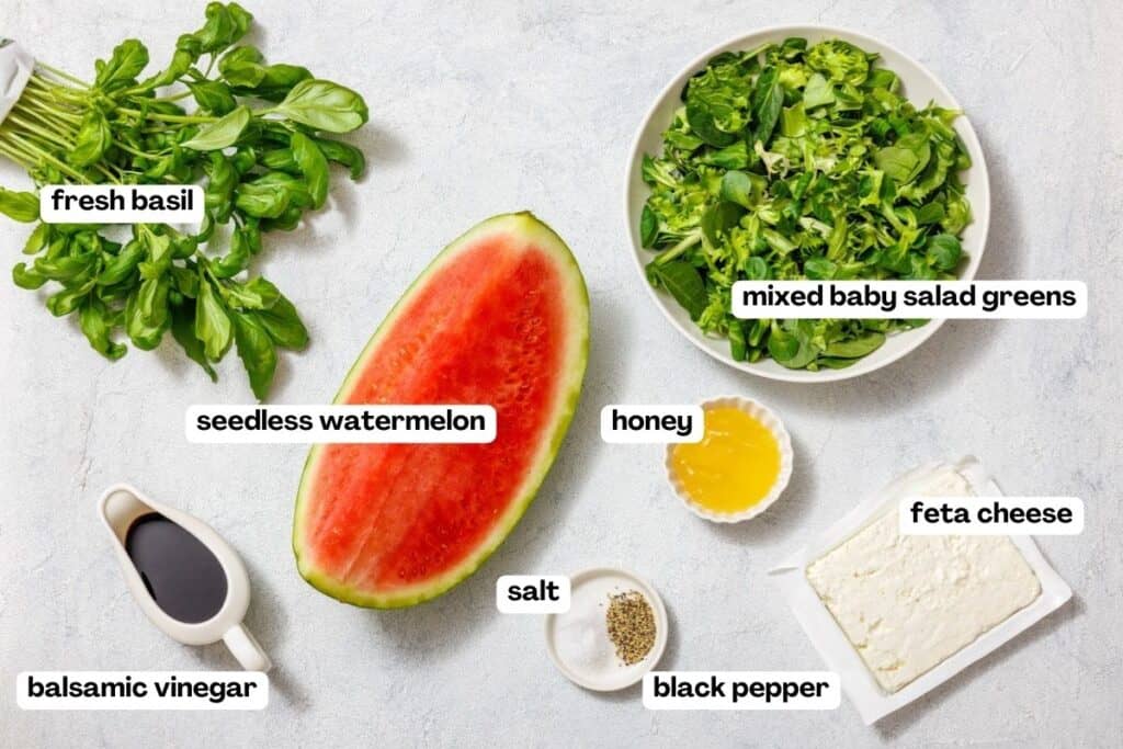 Ingredients for a watermelon salad are laid out: fresh basil, mixed baby salad greens, seedless watermelon, honey, feta cheese, balsamic vinegar, salt, and black pepper.