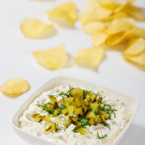 A bowl of creamy Dill Pickle Dip topped with chopped pickles and dill, with scattered potato chips in the background on a white surface.