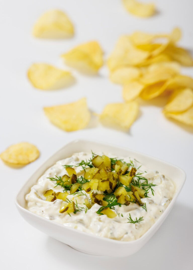 A bowl of creamy Dill Pickle Dip topped with chopped pickles and dill, with scattered potato chips in the background on a white surface.
