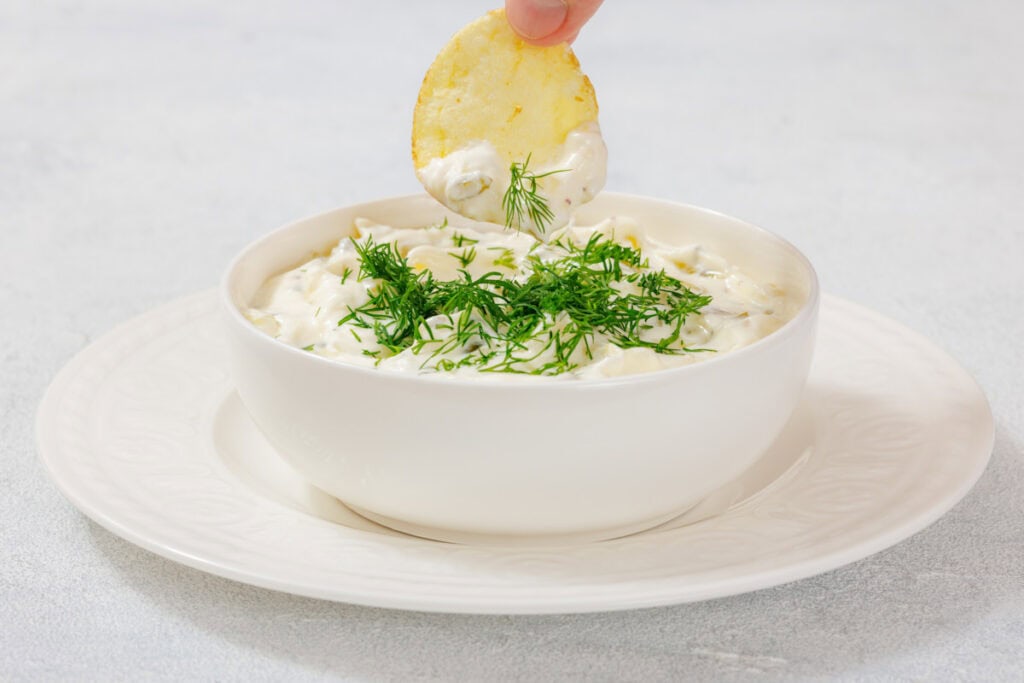"A creamy dill pickle dip garnished with fresh dill, served in a white bowl on a matching white plate, with a hand dipping a potato chip into the dip.