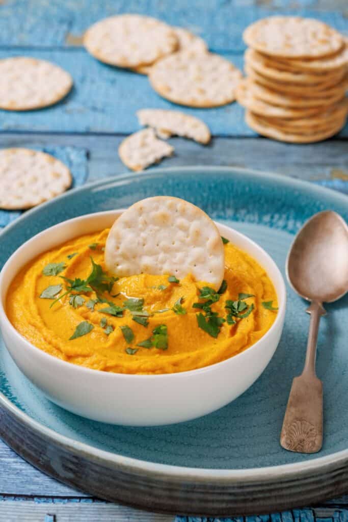A bowl of Roasted Carrot Hummus garnished with chopped herbs and a cracker, placed on a blue plate beside a vintage spoon, surrounded by scattered crackers on a wooden surface.