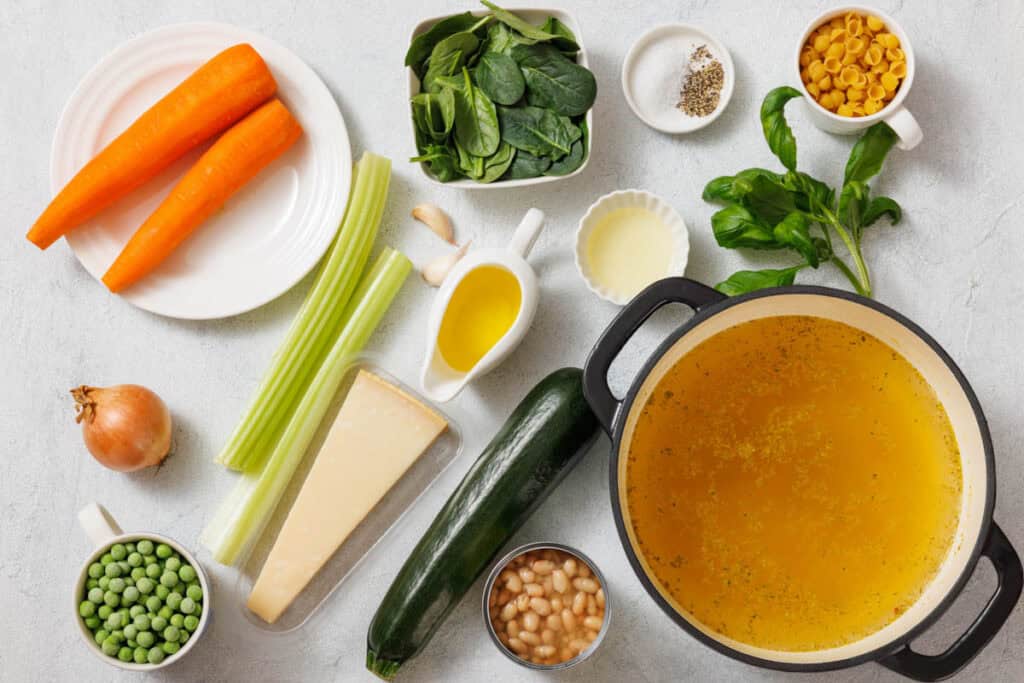 Ingredients for soup on a surface, including carrots, spinach, pasta, celery, olive oil, zucchini, cheese, peas, beans, onion, salt, pepper, and broth in a pot.