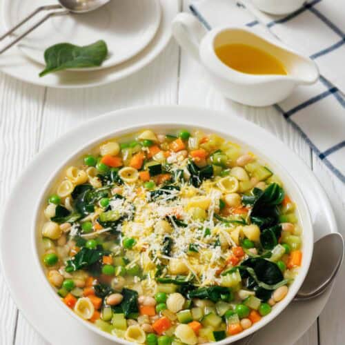 A bowl of Spring Minestrone with pasta, beans, and spinach, topped with grated cheese. A spoon rests beside the bowl. Oil in a gravy boat and salt and pepper shakers are in the background.
