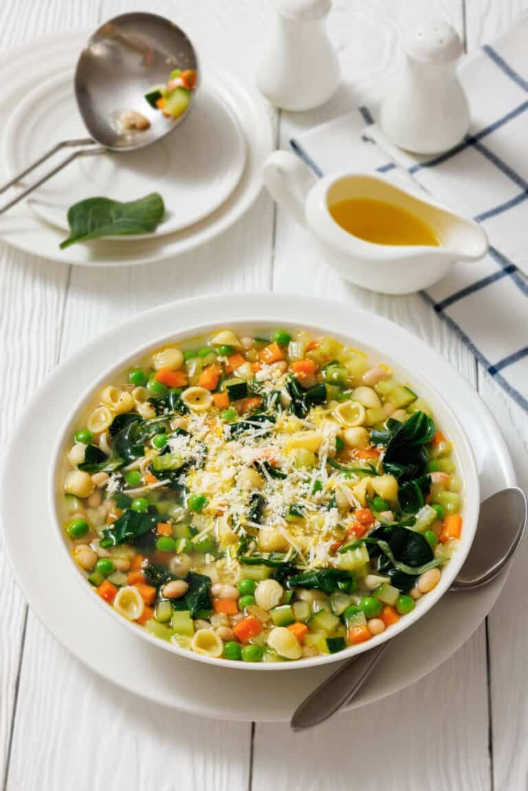 A bowl of Spring Minestrone with pasta, beans, and spinach, topped with grated cheese. A spoon rests beside the bowl. Oil in a gravy boat and salt and pepper shakers are in the background.