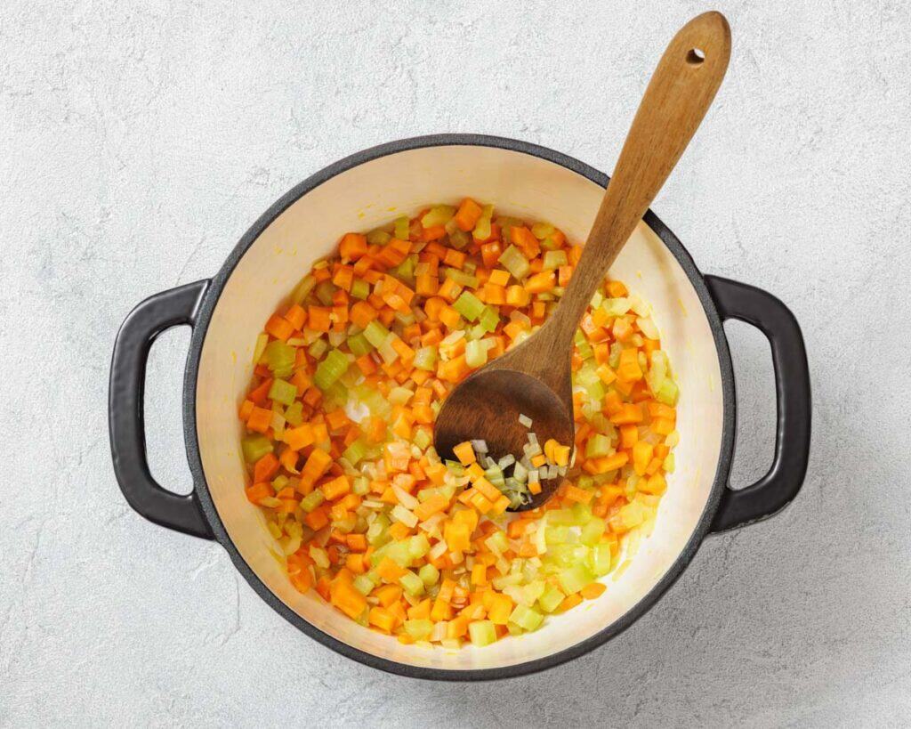 A pot with chopped onions, carrots, and celery being stirred by a wooden spoon.