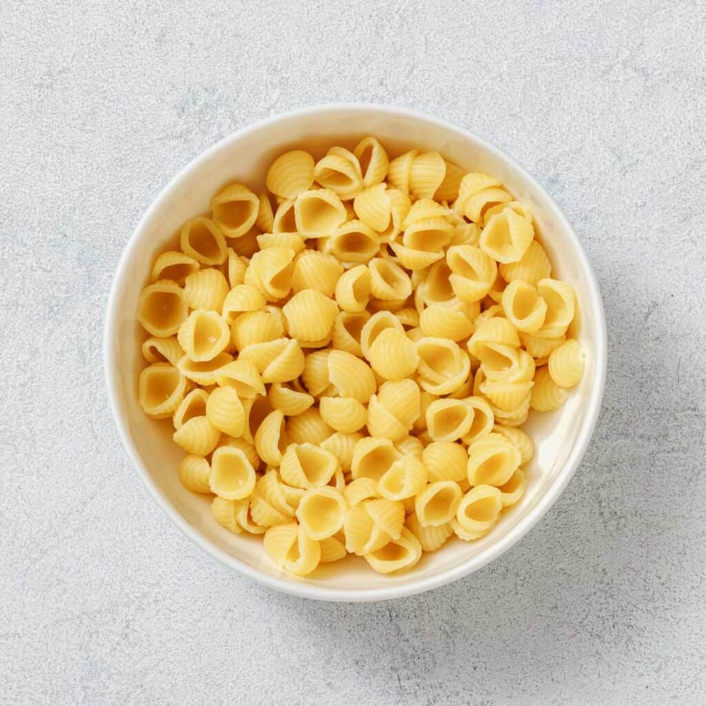 White bowl filled with uncooked, shell-shaped pasta on a light textured surface.