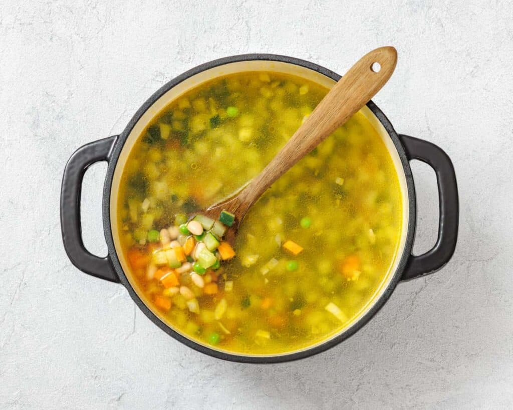 A pot of vegetable soup with a wooden spoon, containing diced carrots, peas, potatoes, and other vegetables, on a light gray background.