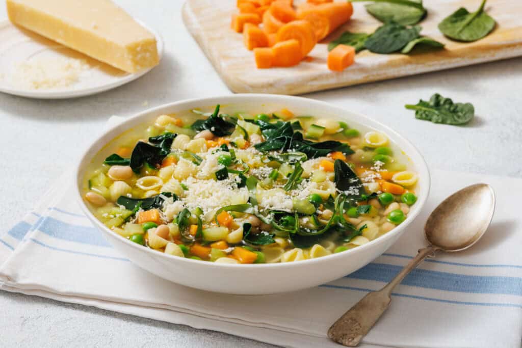 A bowl of Spring Minestrone with pasta and spinach, topped with grated cheese. A spoon rests beside it, and diced carrots and spinach leaves are on a cutting board in the background.