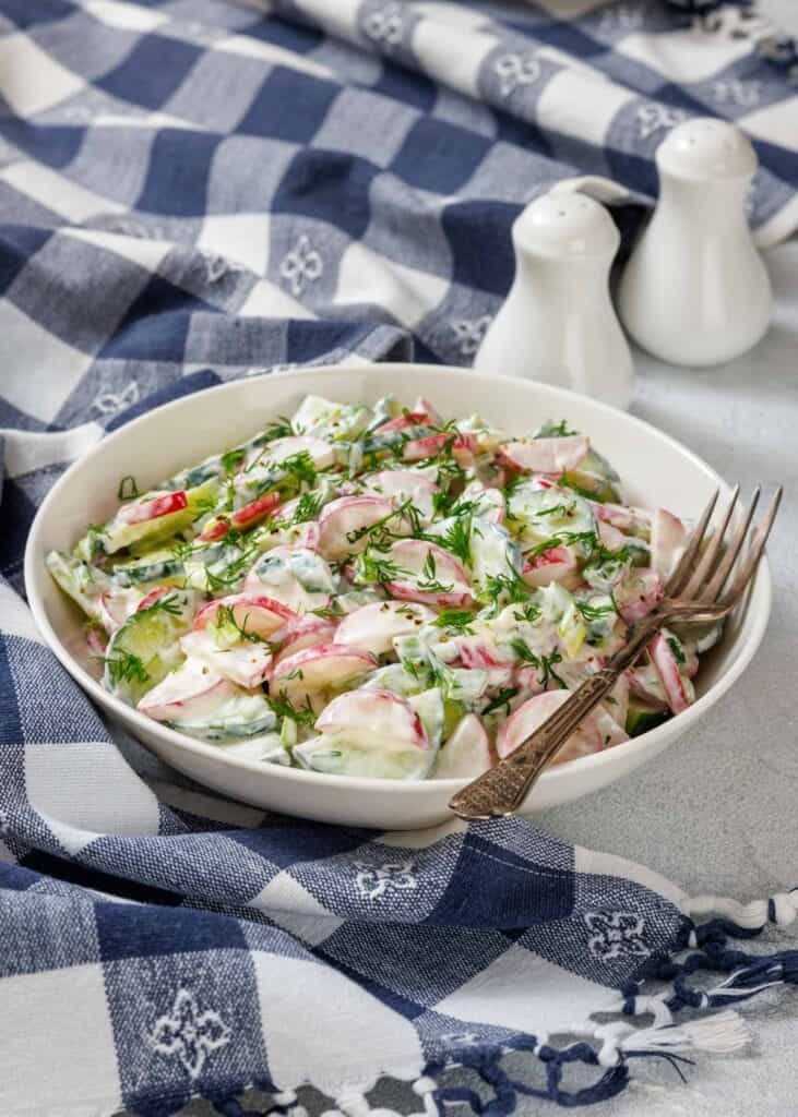 A bowl of creamy vegetable salad with cucumbers, radishes, and herbs on a checkered cloth. Two forks rest on the bowl. Salt and pepper shakers are in the background.