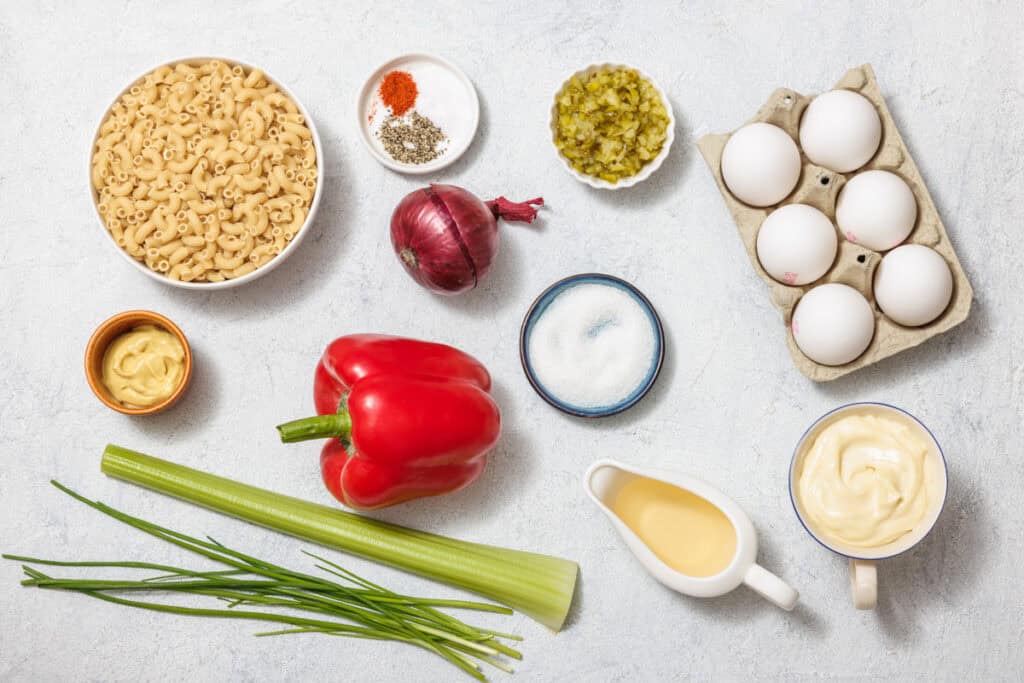 Ingredients for a Deviled Egg Pasta Salad are laid out: macaroni, eggs, red onion, red pepper, celery, chives, mayonnaise, mustard, diced pickles, vinegar, sugar, and seasonings.