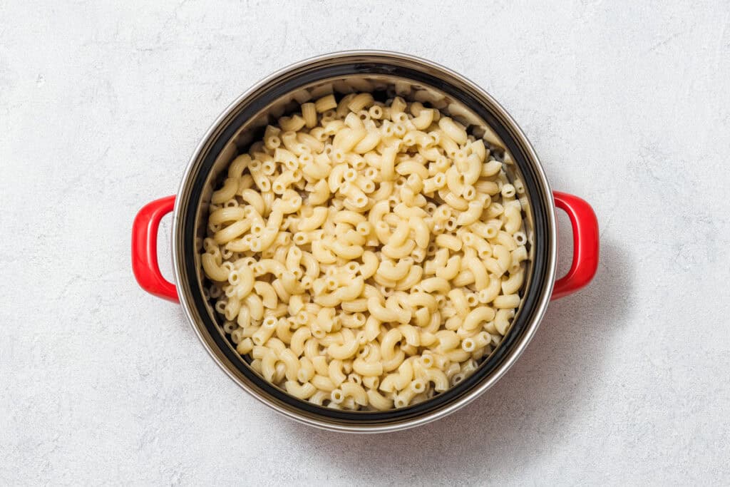 A top-down view of a pot filled with cooked elbow macaroni, featuring red handles, placed on a light-colored textured surface.