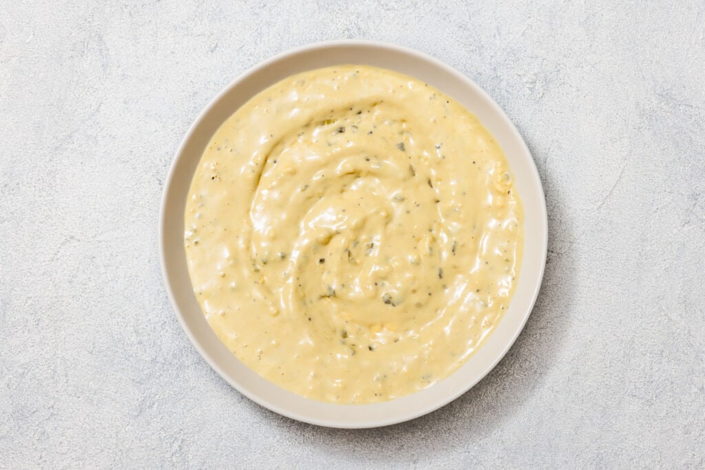 A bowl of creamy, pale yellow sauce with visible herbs, placed on a light textured surface.