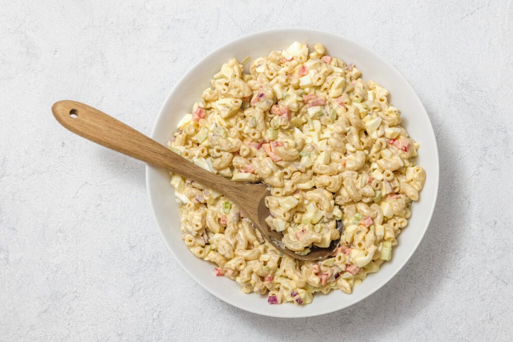 A bowl of macaroni salad with diced vegetables and a wooden spoon on a light surface.
