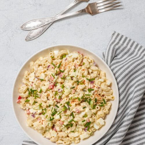 A creamy deviled egg pasta salad served in a white bowl, garnished with chopped chives and paprika, placed on a striped napkin with vintage forks beside it.