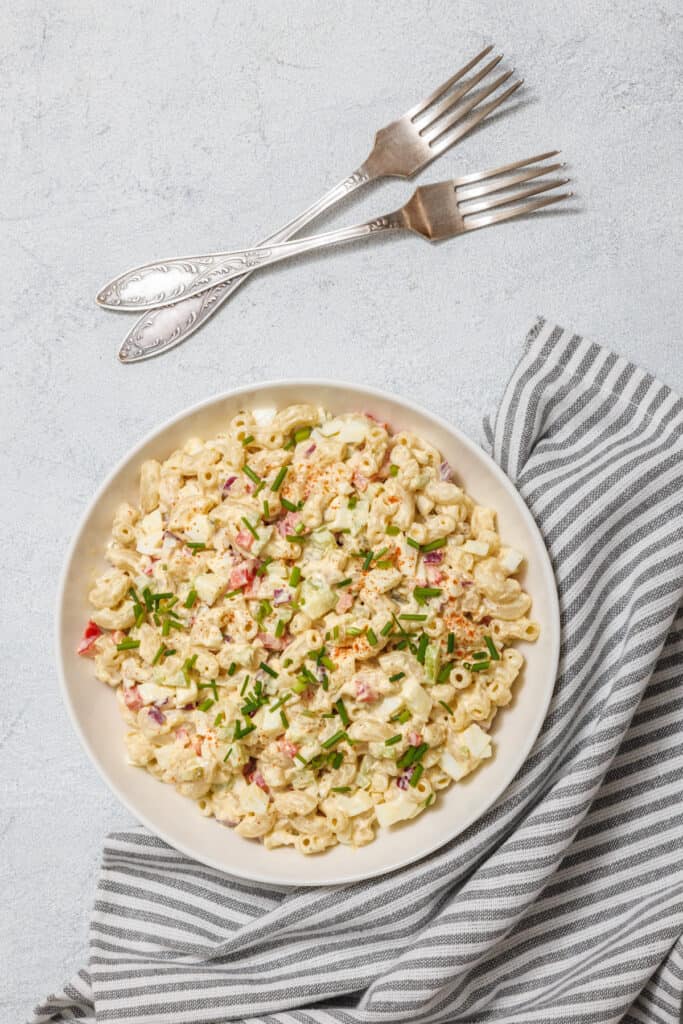 A creamy deviled egg pasta salad served in a white bowl, garnished with chopped chives and paprika, placed on a striped napkin with vintage forks beside it.
