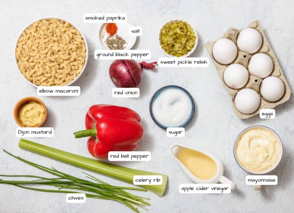 Ingredients for a Deviled Egg Pasta Salad, including elbow macaroni, eggs, red bell pepper, ground black pepper, celery, chives, mayonnaise, and seasonings, arranged on a light surface.