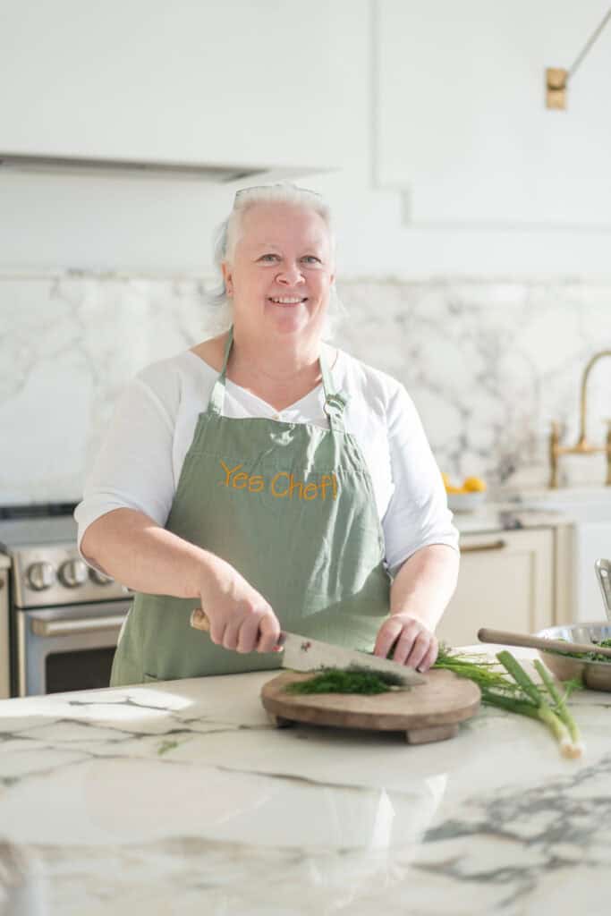 Person in a green apron chopping herbs on a wooden board in a bright kitchen with marble countertops.