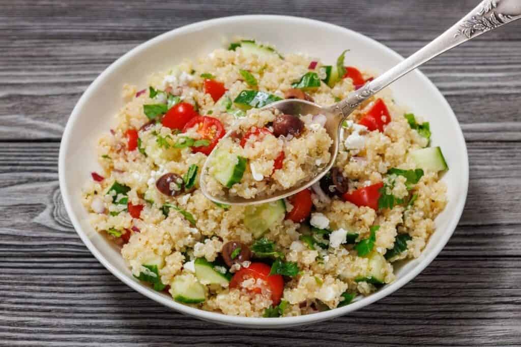 Bowl of quinoa salad with cherry tomatoes, cucumber, olives, feta, and herbs, on a wooden surface, with a spoon lifting a portion.