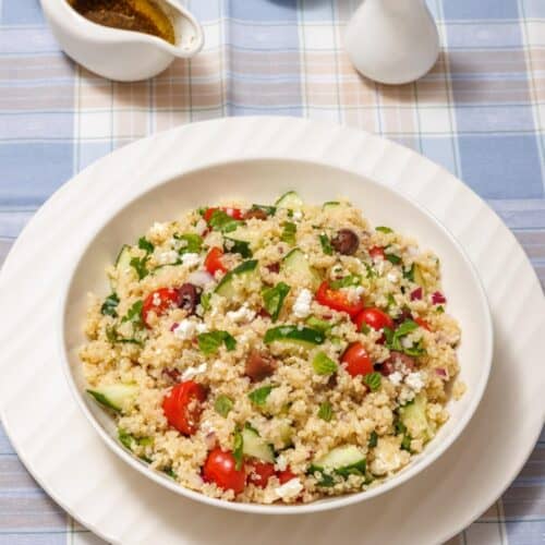 A bowl of quinoa salad with tomatoes, cucumbers, olives, and feta on a checkered tablecloth, accompanied by salt, pepper shakers, and a jug of dressing.