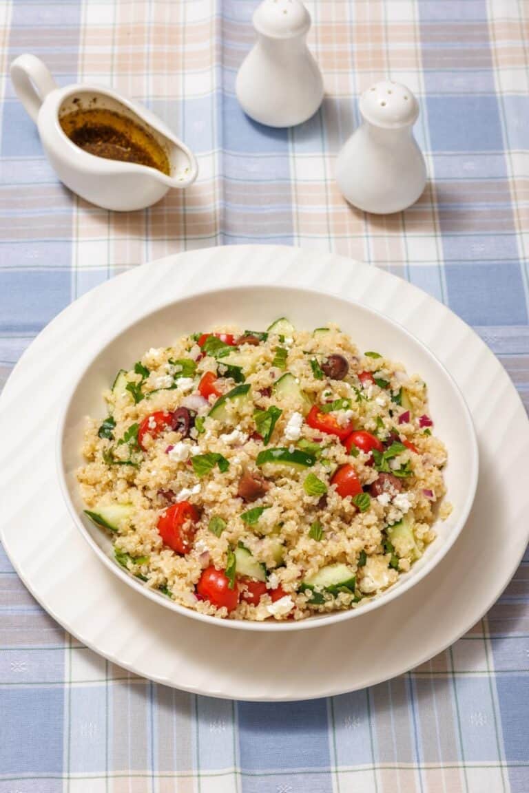 A bowl of quinoa salad with tomatoes, cucumbers, olives, and feta on a checkered tablecloth, accompanied by salt, pepper shakers, and a jug of dressing.
