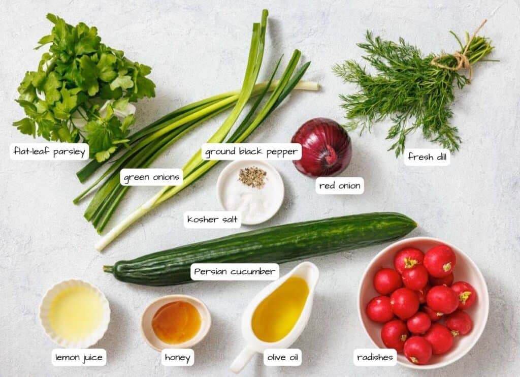 Ingredients laid out for a salad, including flat-leaf parsley, green onions, ground black pepper, red onion, fresh dill, Persian cucumber, kosher salt, lemon juice, honey, olive oil, and radishes.