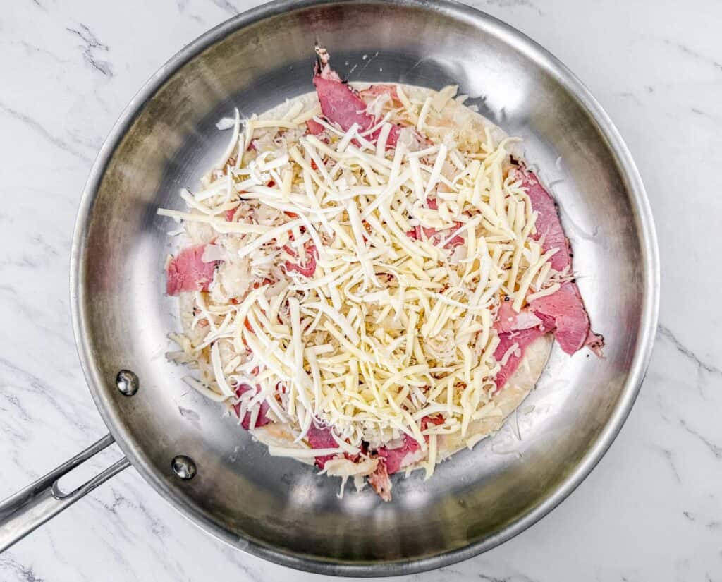 A skillet on a marble surface containing shredded cheese, sliced meat, and sauerkraut.