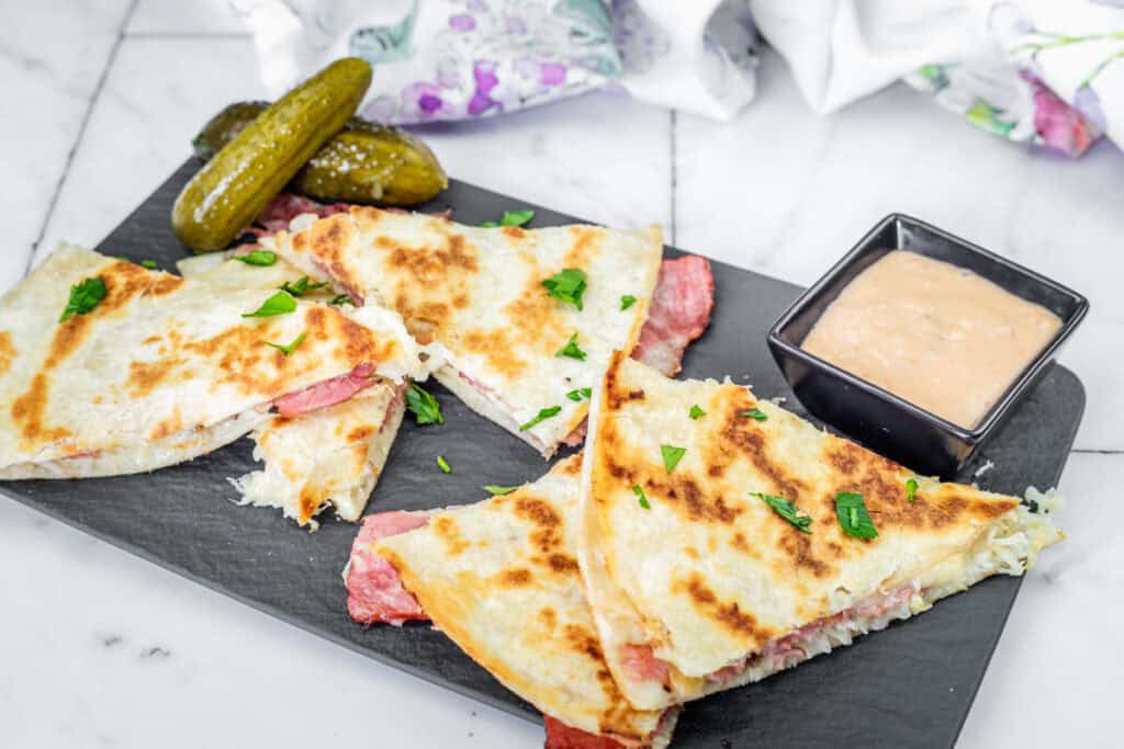 Quesadillas with garnished cilantro, pickles, and a side of dipping sauce on a slate serving board.
