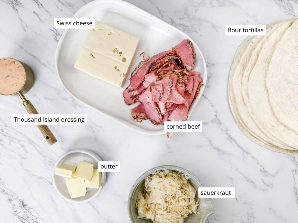 Corned beef, Swiss cheese, sauerkraut, flour tortillas, Thousand Island dressing in a spoon, and cubes of butter arranged on a marble surface.