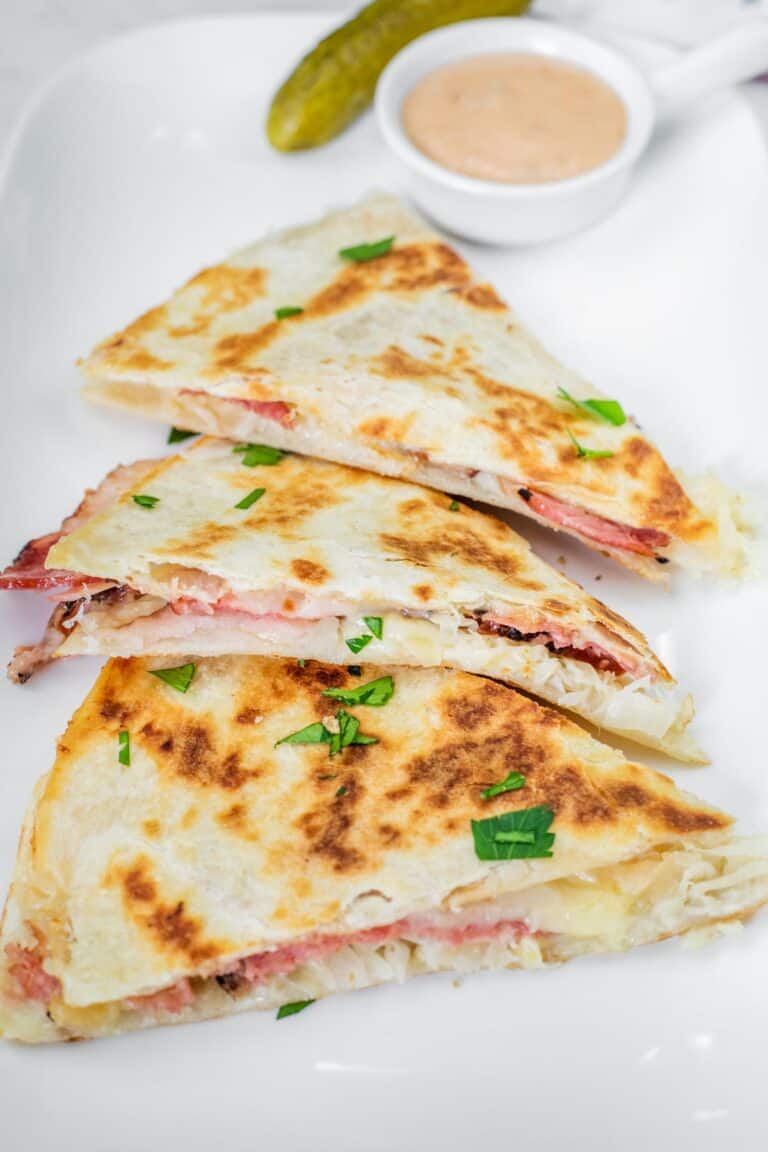 Three sliced quesadillas filled with meat and cheese, garnished with chopped herbs, served on a white plate with a small dish of sauce and a pickle.