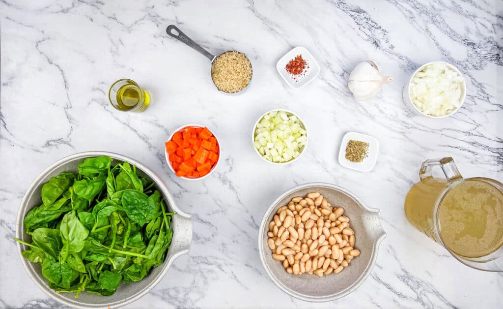 Ingredients on a marble surface: fresh spinach, diced carrots, chopped onions, garlic, olive oil, uncooked rice, broth, white beans, and seasoning.