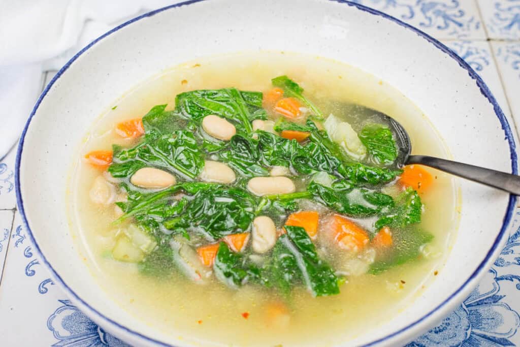 A bowl of Rice and White Bean Soup containing white beans, kale, carrots, and other vegetables, with a spoon on the side.