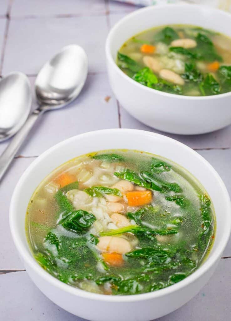Two white bowls of Rice and White Bean Soup on a tiled surface with two metal spoons nearby.