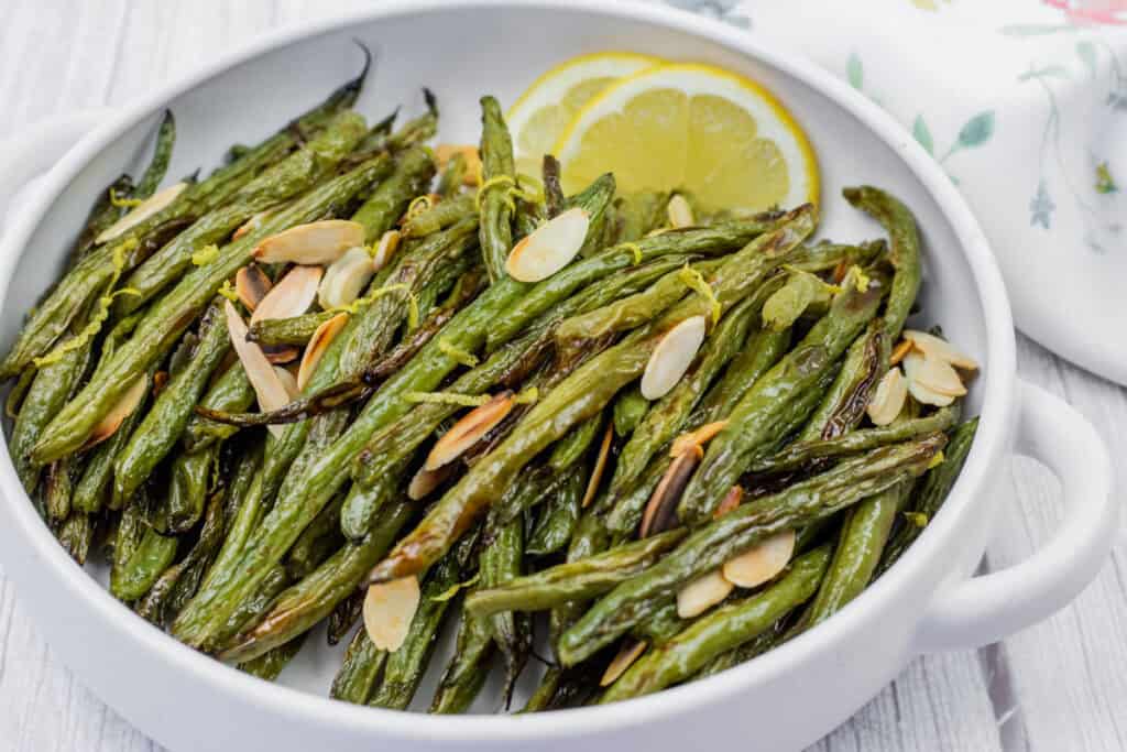 A white bowl filled with roasted green beans topped with sliced almonds, alongside lemon slices, on a floral napkin.