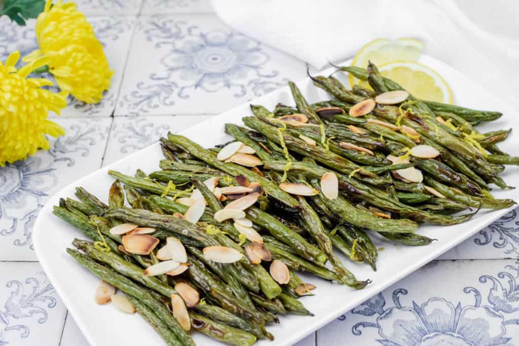 A platter of roasted green beans topped with sliced almonds, placed on a patterned surface. Yellow flowers and lemon slices are visible in the background.