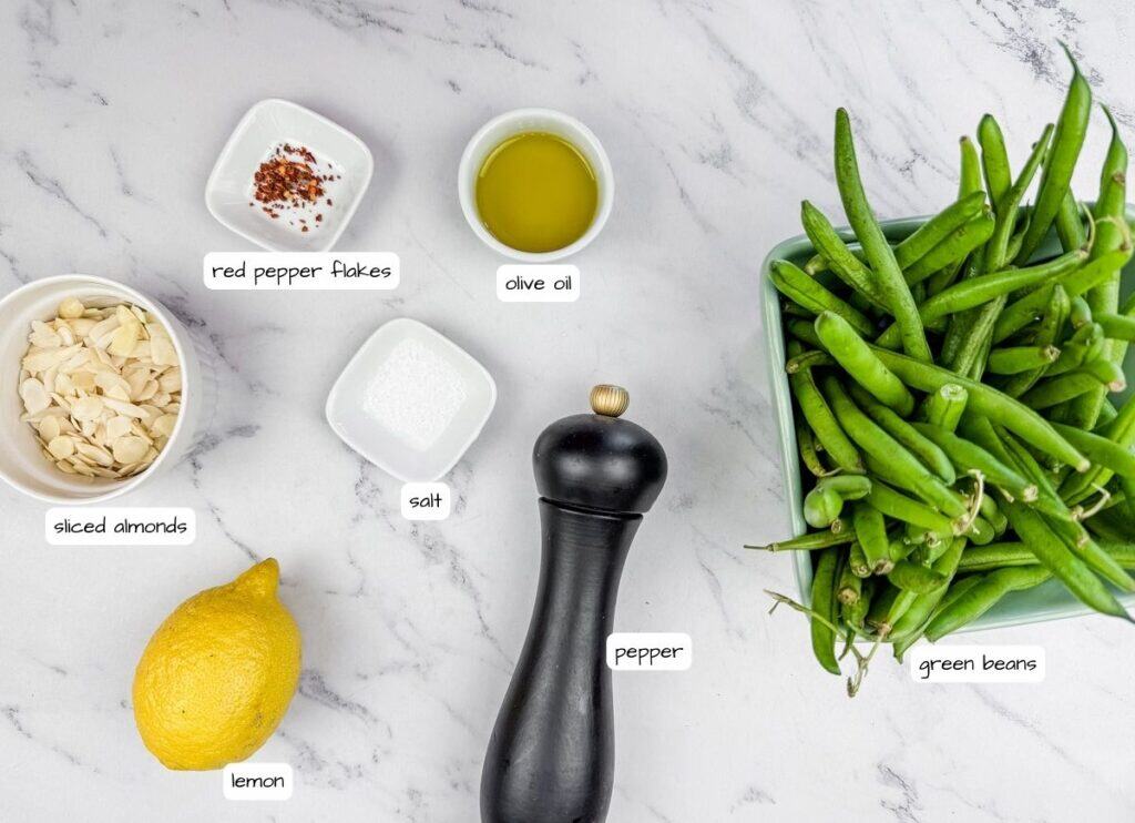 Ingredients on a marble surface: sliced almonds, red pepper flakes, olive oil, salt, whole pepper grinder, fresh green beans, and a lemon.