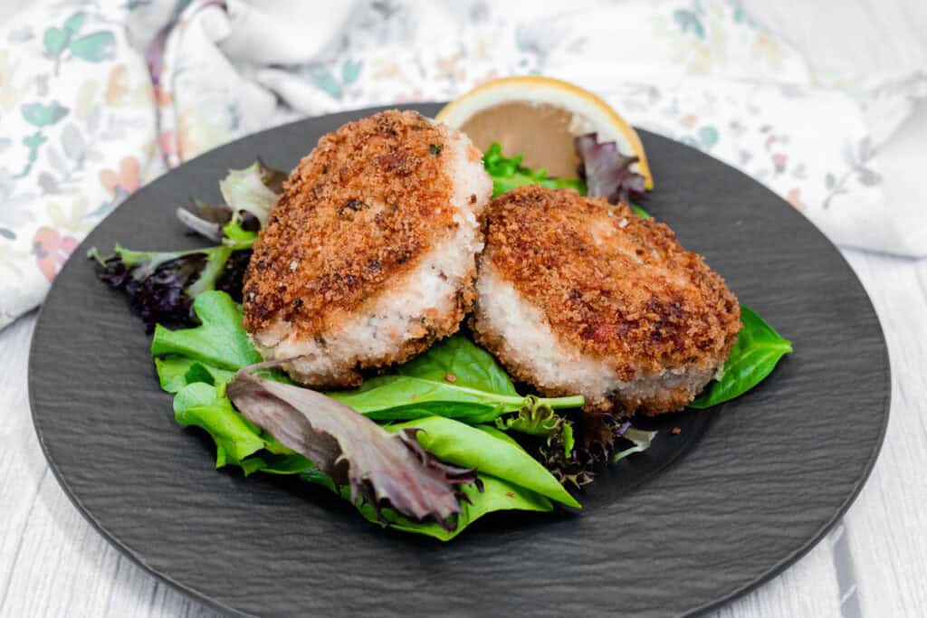 Two Salmon Cakes on a bed of mixed greens, garnished with a lemon wedge, served on a black plate.