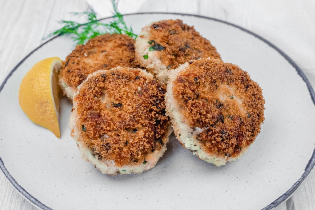 Four Salmon Cakes are on a plate with a lemon wedge and a sprig of dill.