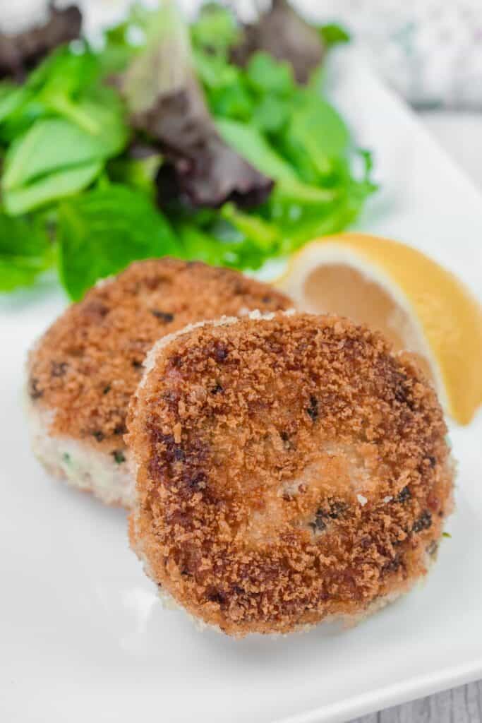 Two Salmon Cakes with a wedge of lemon and mixed greens on a white plate.