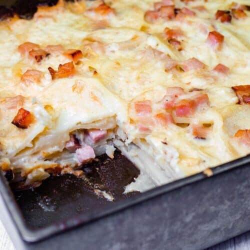 A Scalloped Potatoes with Ham and Cheese featuring layers of sliced potatoes, ham, and melted cheese with a portion removed, revealing the ingredients.