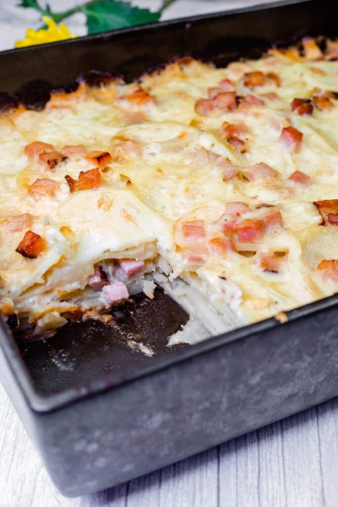 A Scalloped Potatoes with Ham and Cheese featuring layers of sliced potatoes, ham, and melted cheese with a portion removed, revealing the ingredients.