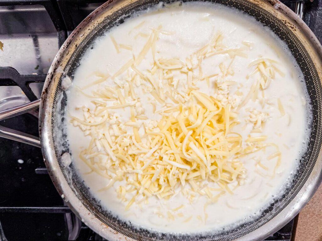 Shredded cheese melting in a frying pan filled with creamy white sauce on a stovetop.