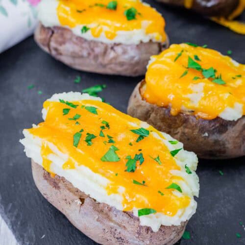 Shepherd’s Pie Baked Potatoes topped with creamy mashed potatoes, melted cheddar cheese, and fresh parsley, served on a black slate board.