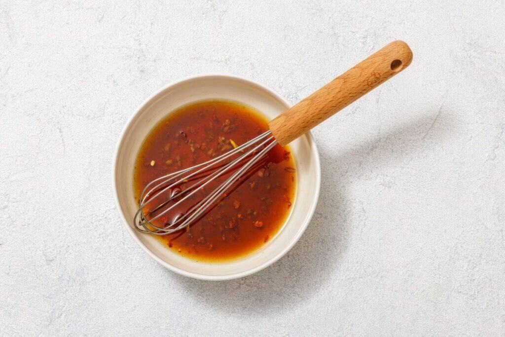 A white bowl filled with a reddish-brown liquid and small solid ingredients, with a whisk resting inside, placed on a light-colored surface.