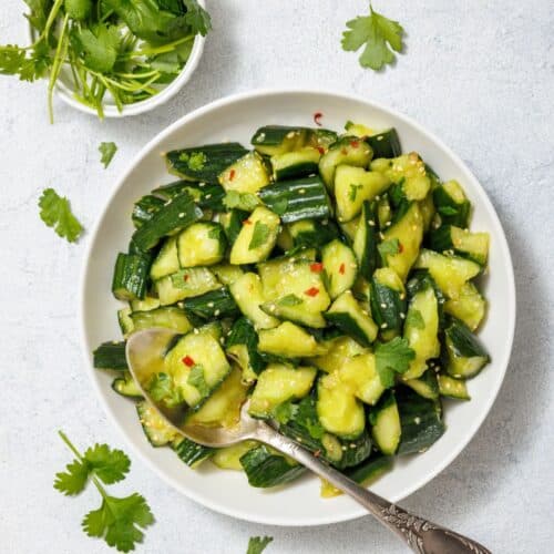 A bowl of chopped cucumber salad with red pepper flakes and cilantro, accompanied by a silver spoon. A small bowl of fresh cilantro is placed next to the salad.