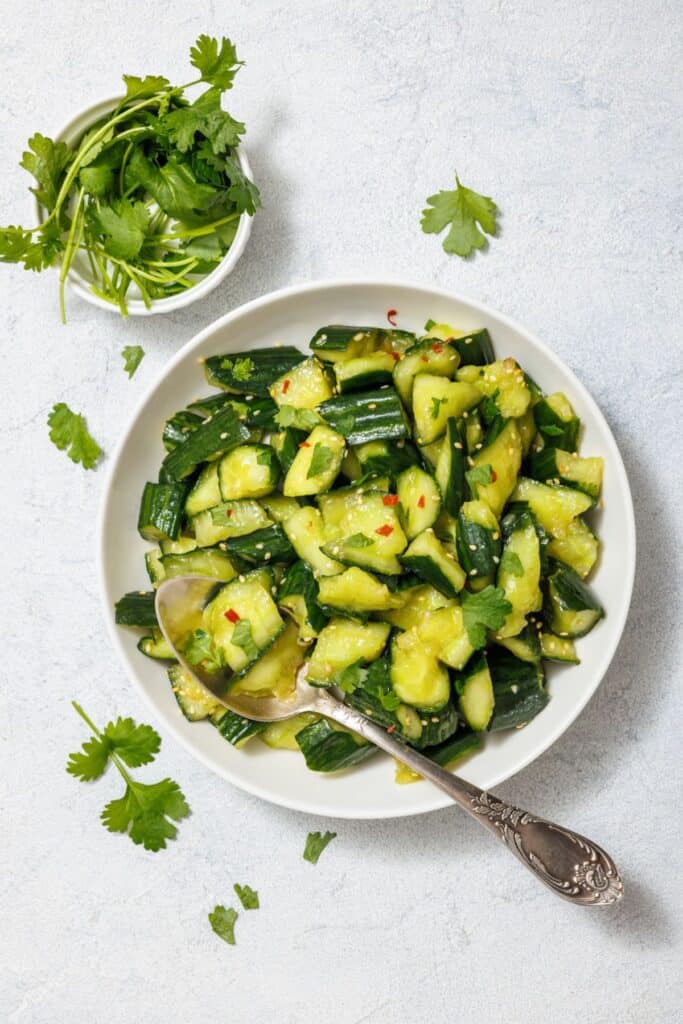 A bowl of chopped cucumber salad with red pepper flakes and cilantro, accompanied by a silver spoon. A small bowl of fresh cilantro is placed next to the salad.