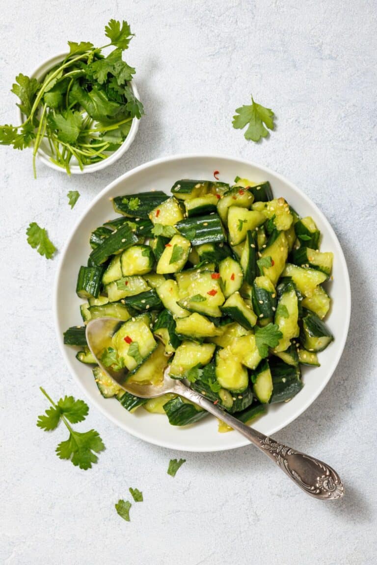 A bowl of chopped cucumber salad with red pepper flakes and cilantro, accompanied by a silver spoon. A small bowl of fresh cilantro is placed next to the salad.