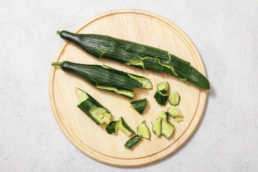 Two partially peeled cucumbers on a wooden cutting board, with several pieces chopped into chunks nearby.
