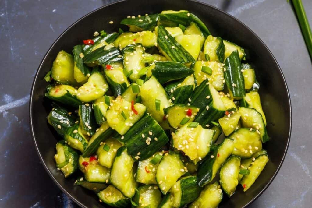 A bowl of chopped cucumbers mixed with sesame seeds, chopped chives, and red pepper flakes.