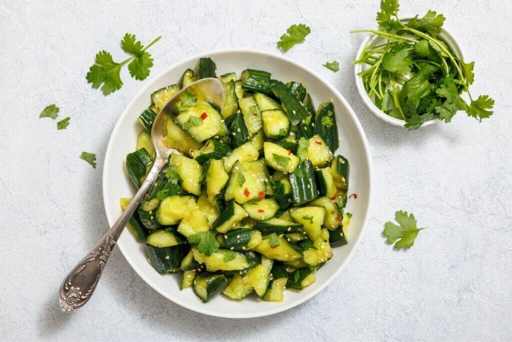 A plate of cucumber salad with chili flakes and cilantro, accompanied by a spoon. A small bowl of cilantro is on the side, with scattered cilantro leaves on the table.