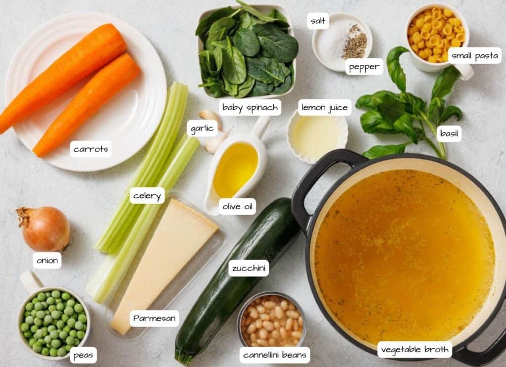 Vegetable broth in a pot surrounded by carrots, celery, onion, peas, spinach, pasta, zucchini, cheese, beans, garlic, olive oil, lemon juice, basil, salt, and pepper on a gray surface.